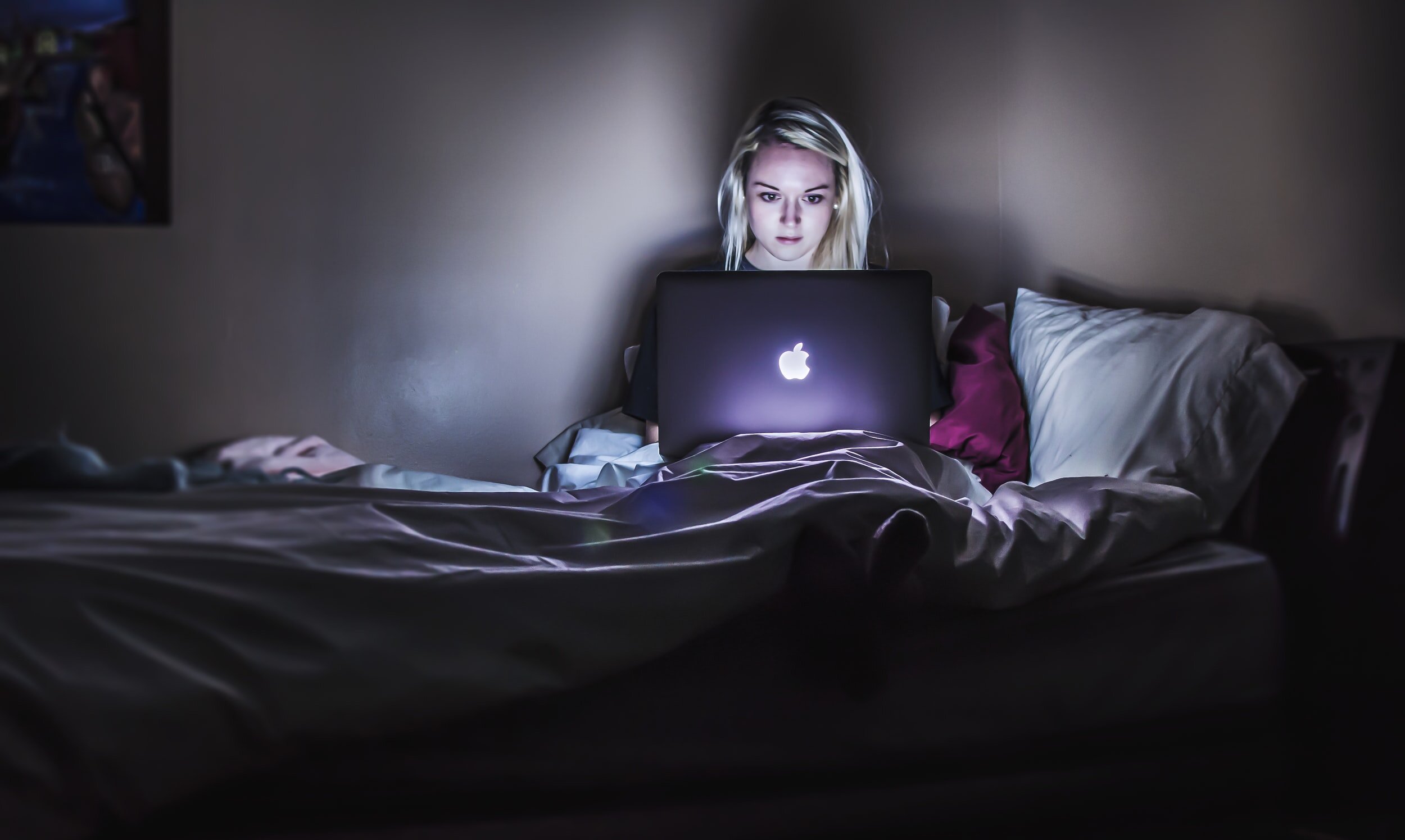 A woman sitting on her bed using her laptop, the screen is illuminating the room