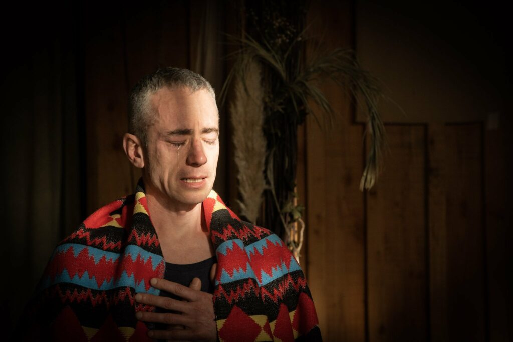 James O'Halloran, eyes closed, and a tear running down his cheek, wearing a colorful patterned blanket and holding his hand over his heart, conveying deep emotion.