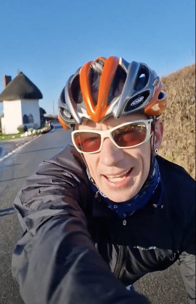 James O'Halloran cycling on a road, wearing a helmet and sunglasses, smiling at the camera.