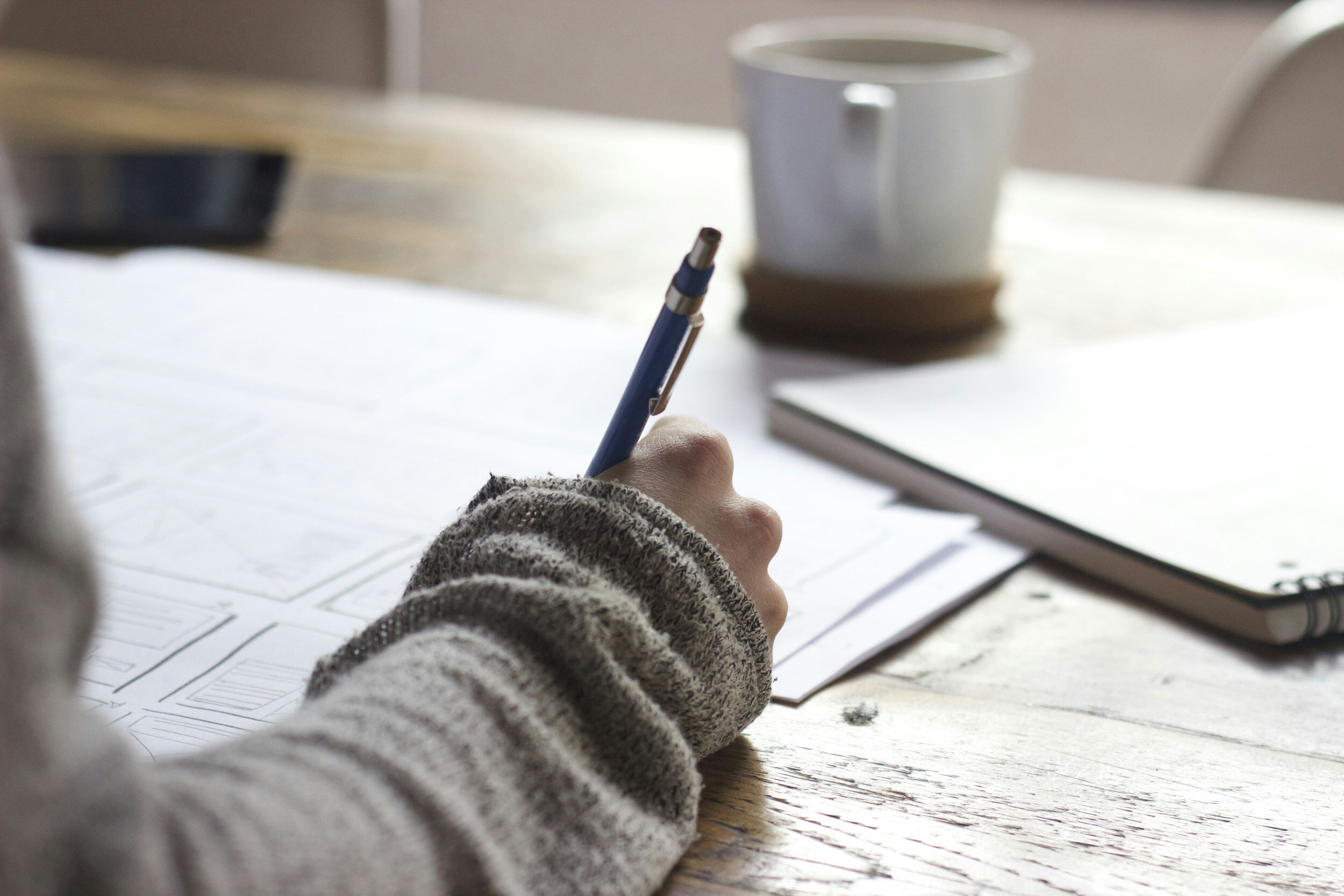 A person writing on paper at a wooden desk with a mug in the background