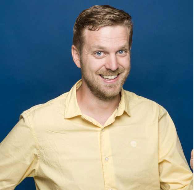 A smiling man wearing a yellow shirt against a blue background.