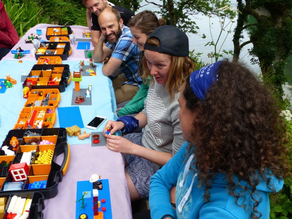 A photo of 5 adults sitting at a table outside. There is lego on the table and smiles on the faces as they work something out together.
