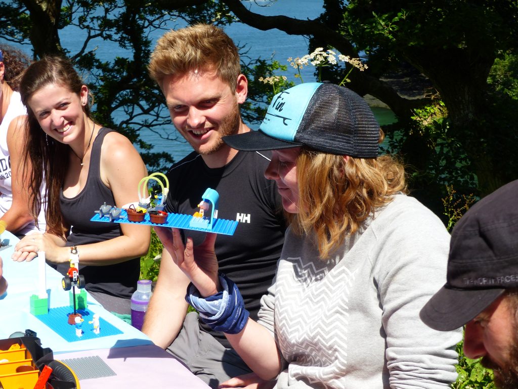 A photo of 3 adults sitting at a table outside. There is lego on the table and smiles on the faces as they work something out together.